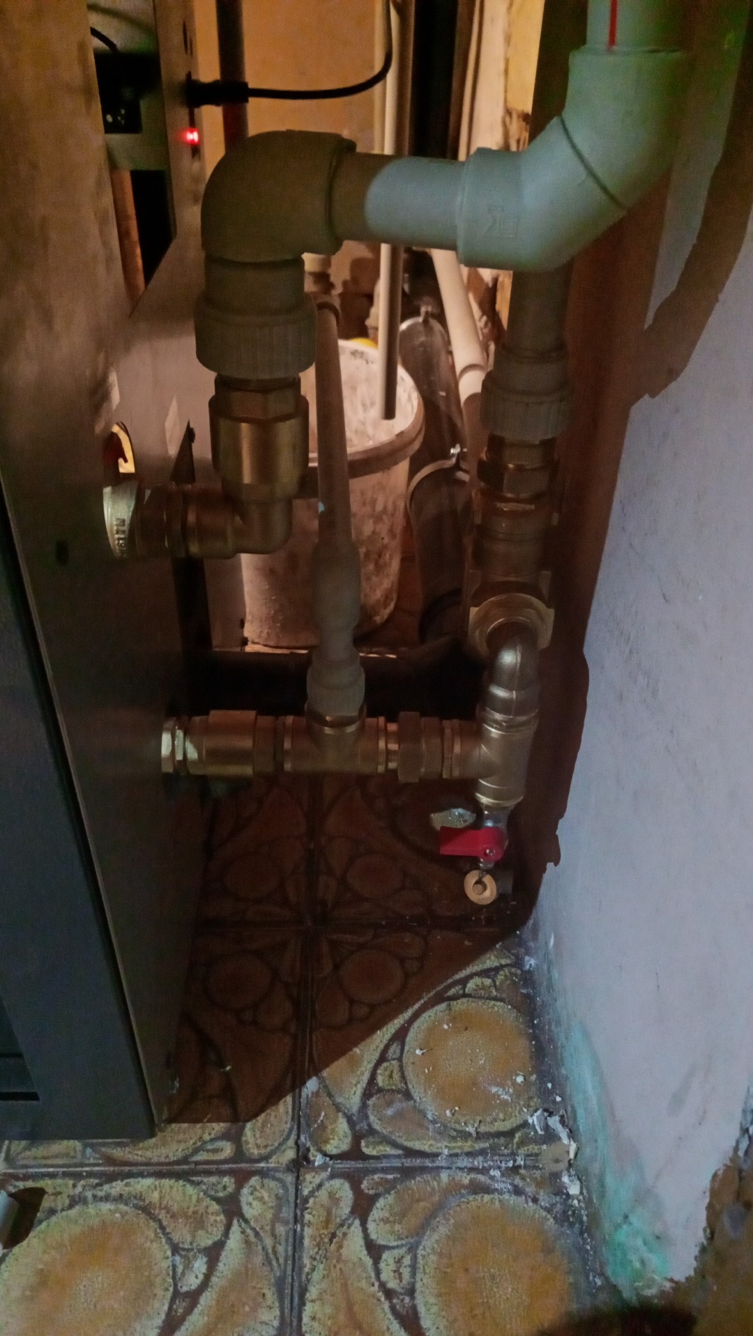 Fragment instalacji hydraulicznej z mosiężnymi złączkami, rurami PCV i zaworem kulowym z czerwoną rączką, widoczne płytki podłogowe z wzorem i fragment ściany z zaciekami.