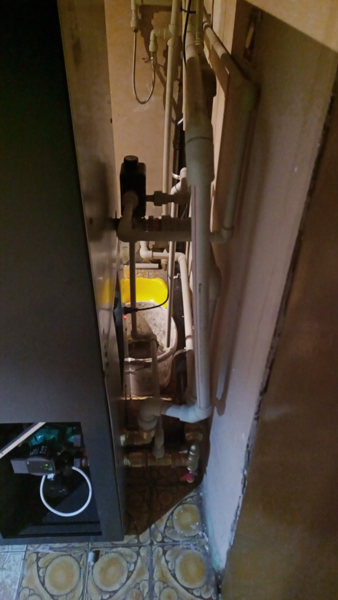 Fragment instalacji hydraulicznej z widocznymi rurami PCV, zaworami i elementami sterującymi w kotłowni, z żółtym elementem w tle i wzorzystą podłogą z płytek.