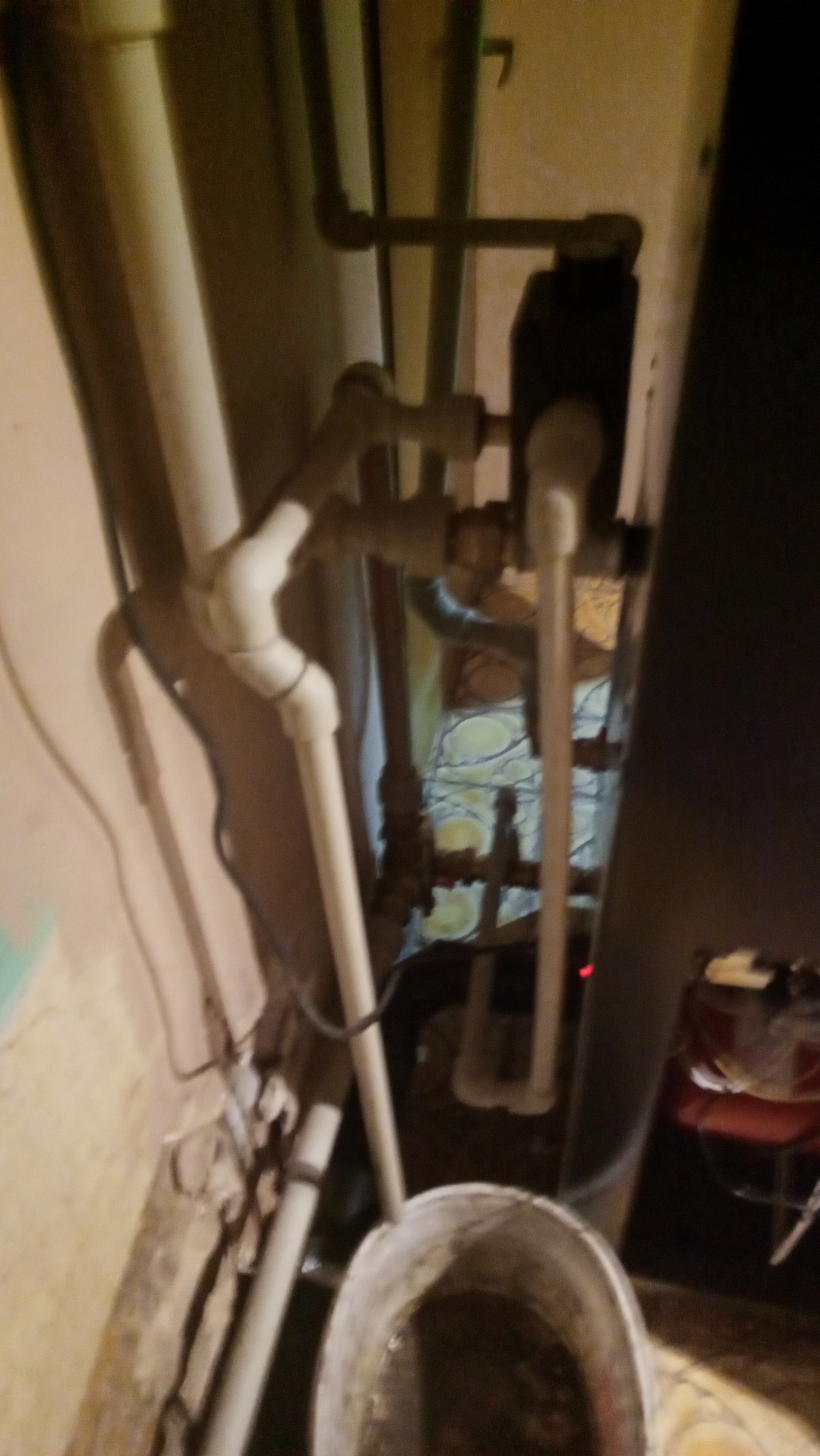 Widok z dołu na instalację hydrauliczną z rurami PCV, z widocznym wiaderkiem zanieczyszczonej wody, w pomieszczeniu o słabym oświetleniu.
