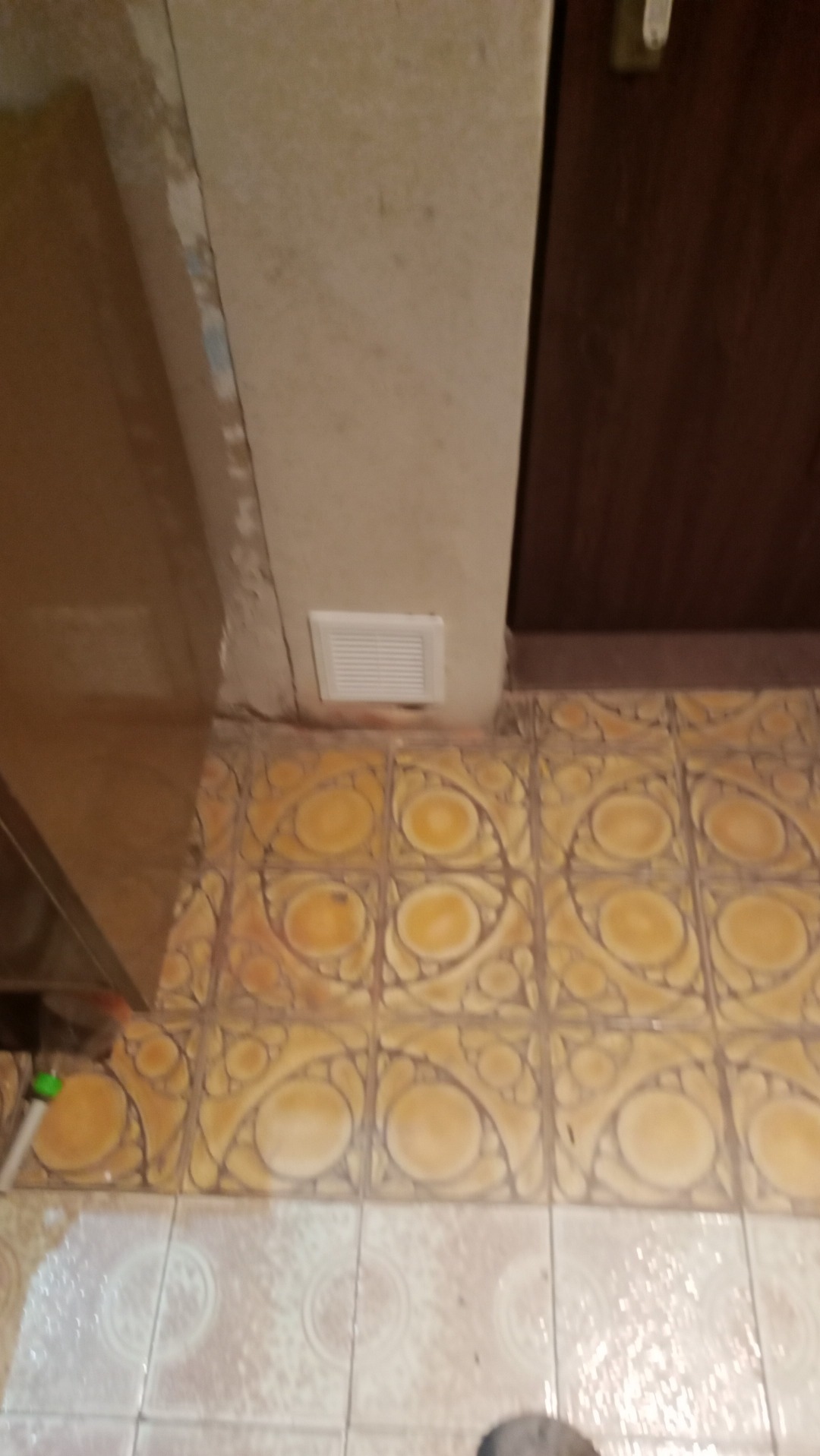 Fragment łazienki z żółtymi płytkami podłogowymi w okrągłe wzory, biała kratka wentylacyjna u dołu ściany, brudne fugi i ślady po zalaniu.