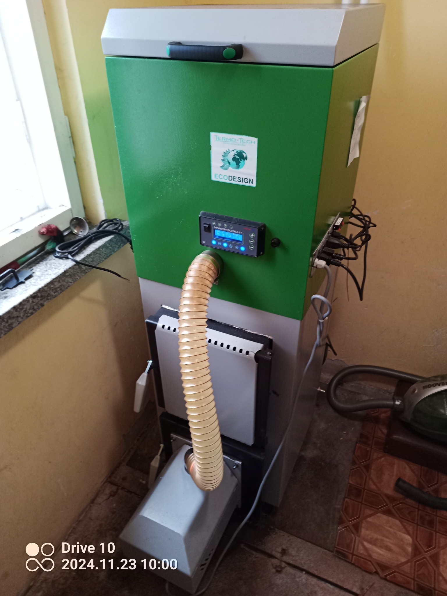 Zielono-szary piec na paliwo stałe z podłączonym wężem i panelem sterowania, zainstalowany w Toruniu, widoczne okno i narzędzia w tle.