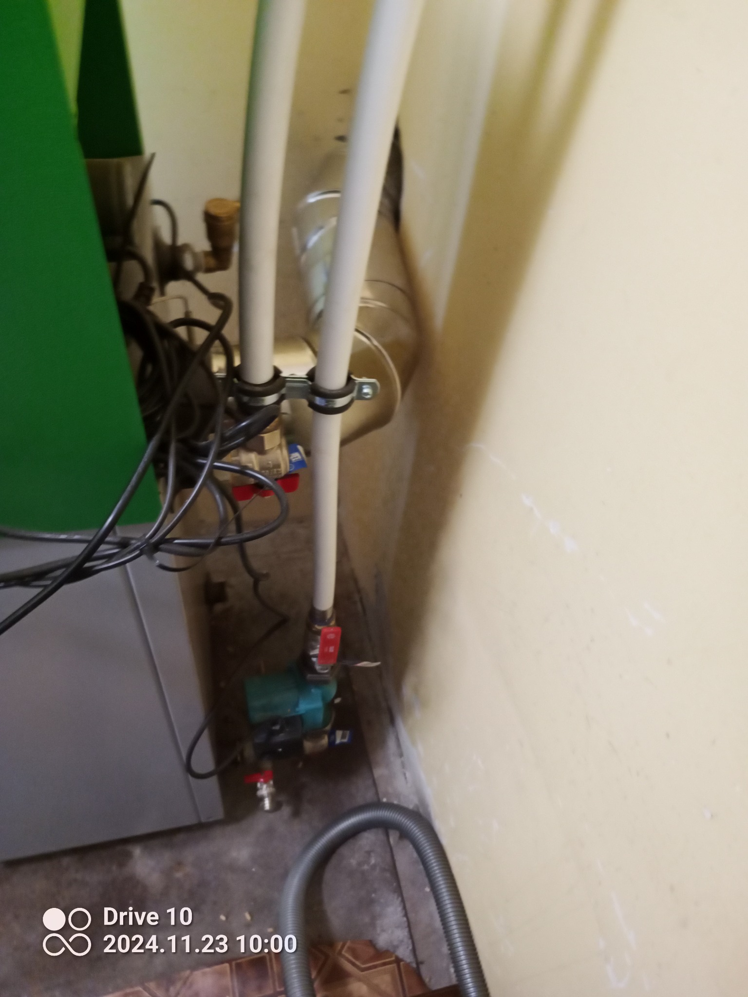 Fragment instalacji grzewczej z widoczną pompą obiegową, rurami z zaworami i przewodami elektrycznymi, umieszczony w narożniku przy beżowej ścianie.