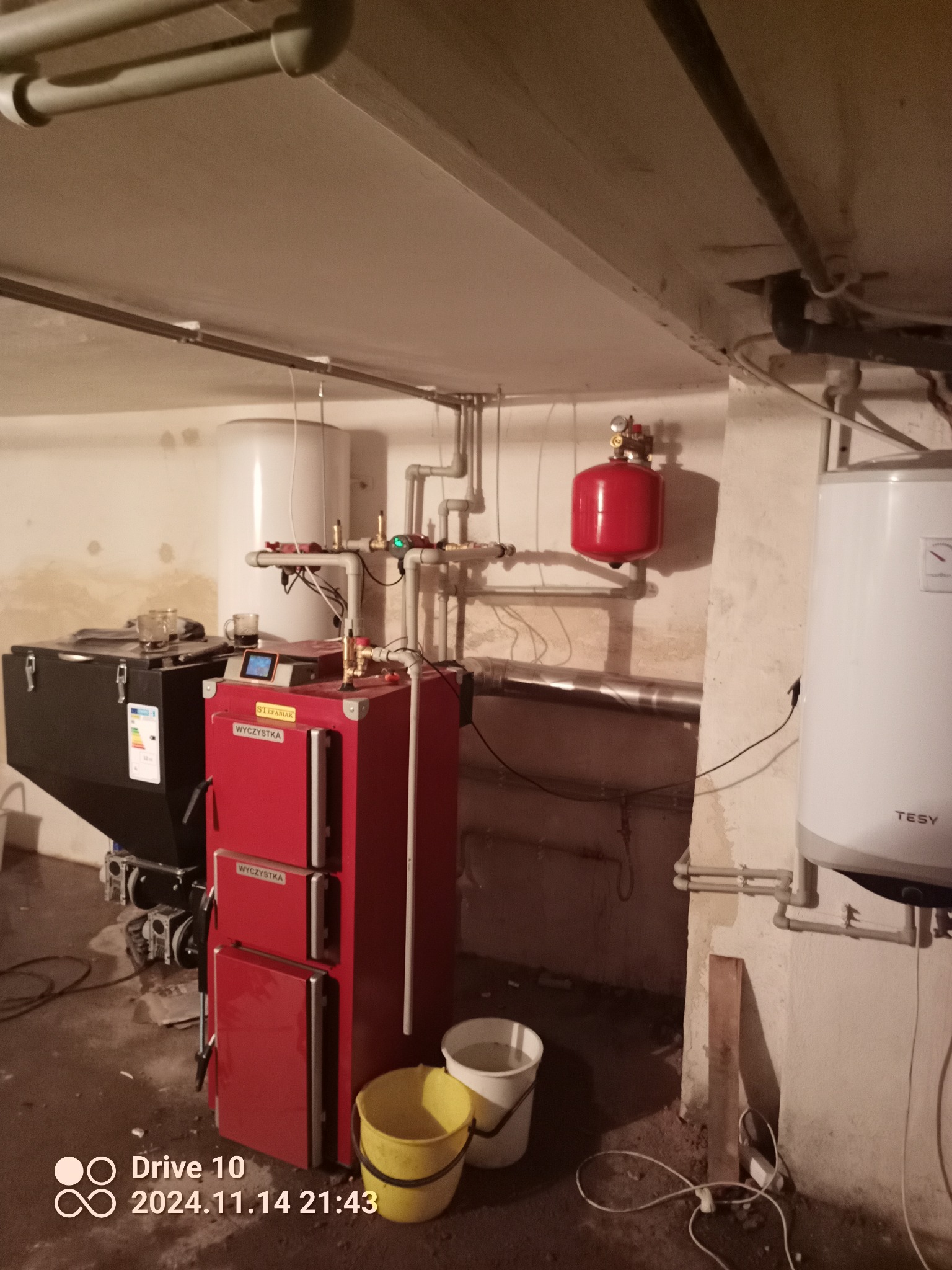 Czerwony piec na paliwo stałe z podajnikiem, widoczne rury instalacji centralnego ogrzewania, zbiornik wyrównawczy oraz dwa wiadra na podłodze w piwnicy.