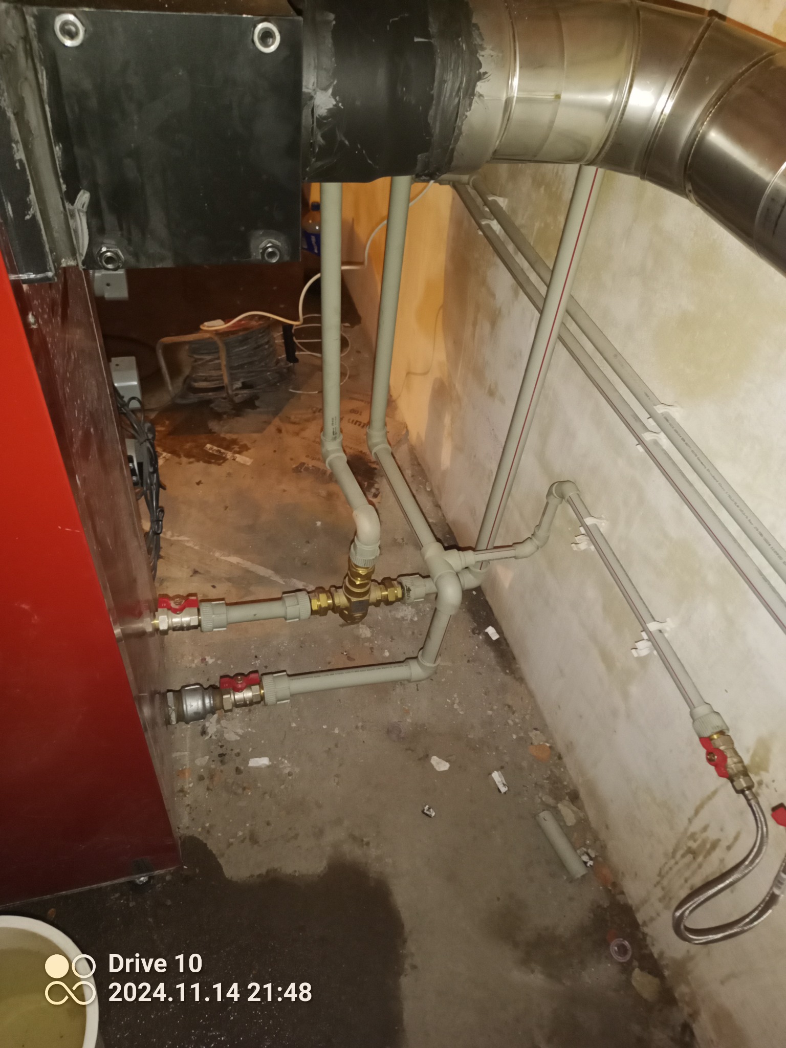 Fragment kotłowni z widocznym piecem na paliwo stałe, rurami instalacji grzewczej z zaworami, elementami wentylacji i wilgotną podłogą.