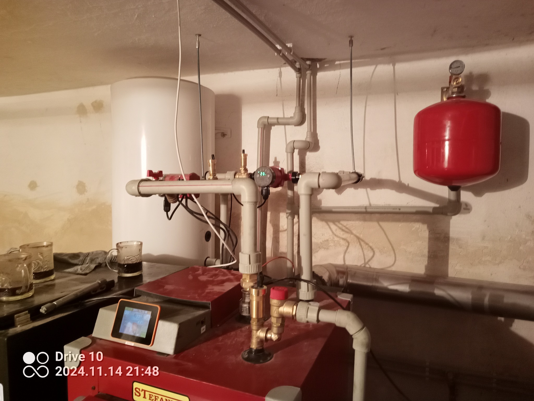 Kotłownia z piecem na paliwo stałe, zbiornikiem wody, rurami i naczyniem przeponowym w kolorze czerwonym, widoczne trzy szklanki z ciemnym płynem i niedbale rzucona ścierka.