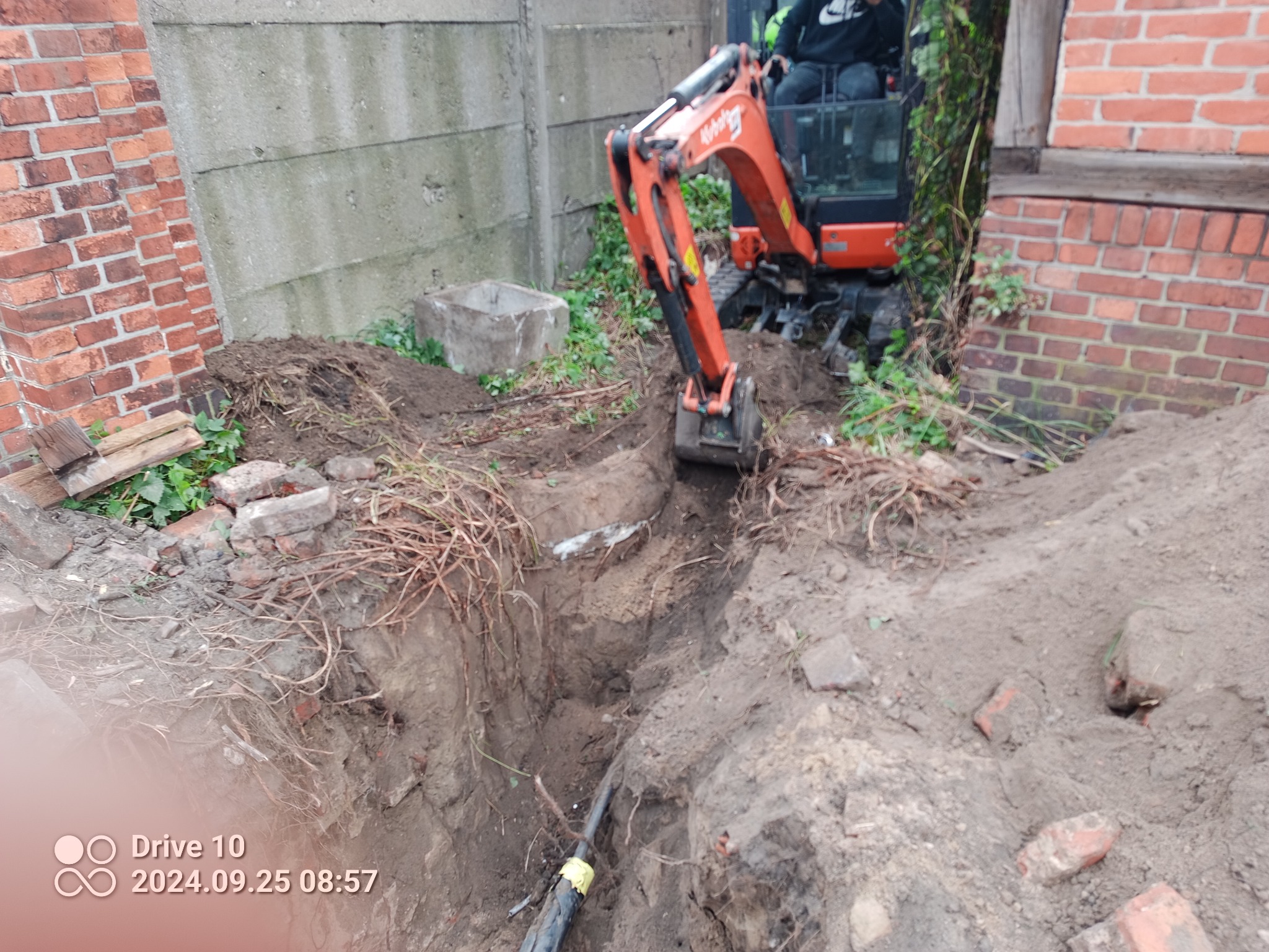 Pomarańczowa minikoparka Kubota pracuje w wykopie ziemnym, widoczne kable w żółtej izolacji, ceglany mur i betonowy płot w tle.