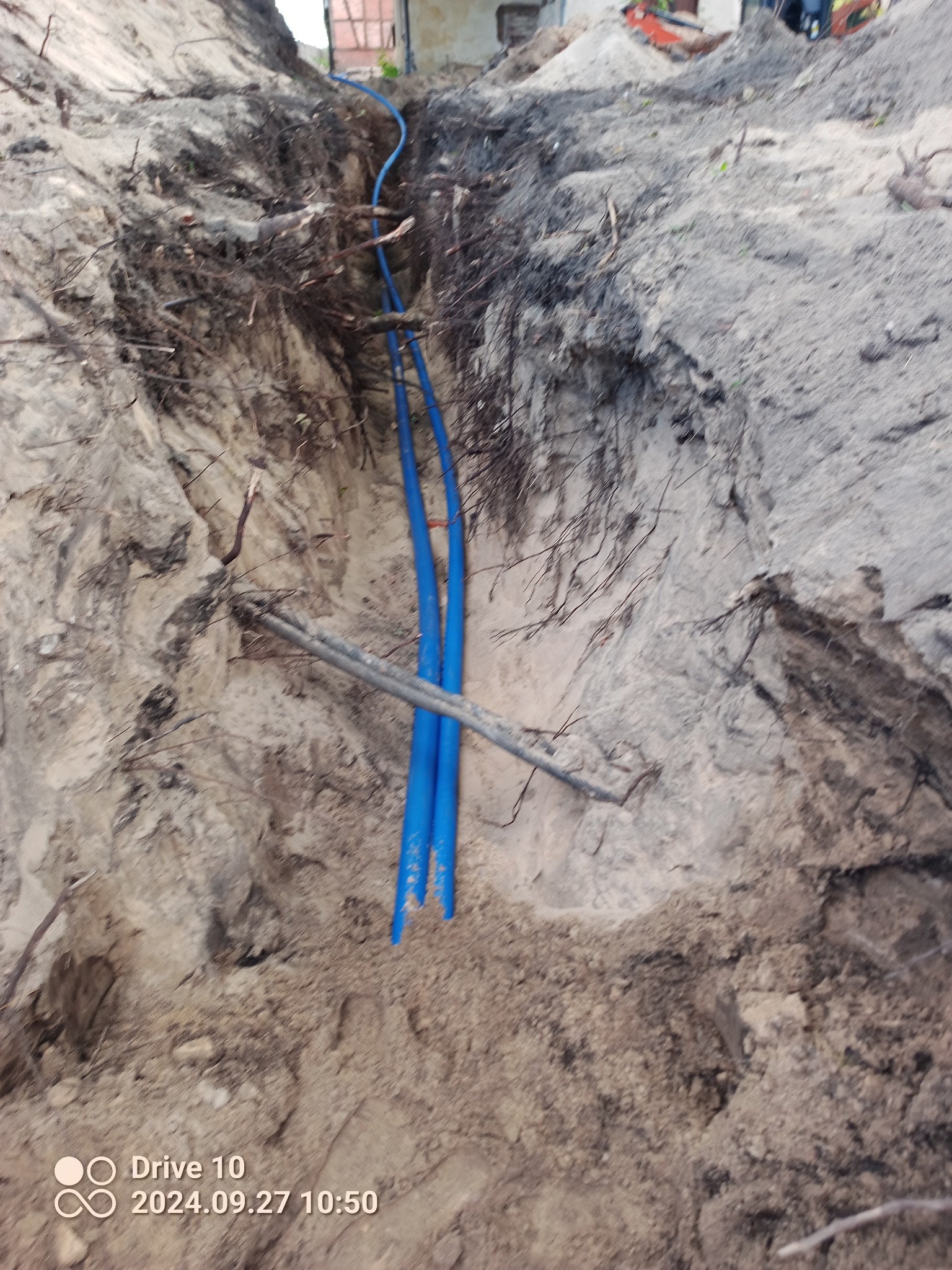 Wykop z widocznymi trzema niebieskimi rurami oraz szarym przewodem, prawdopodobnie podczas układania w ziemi.