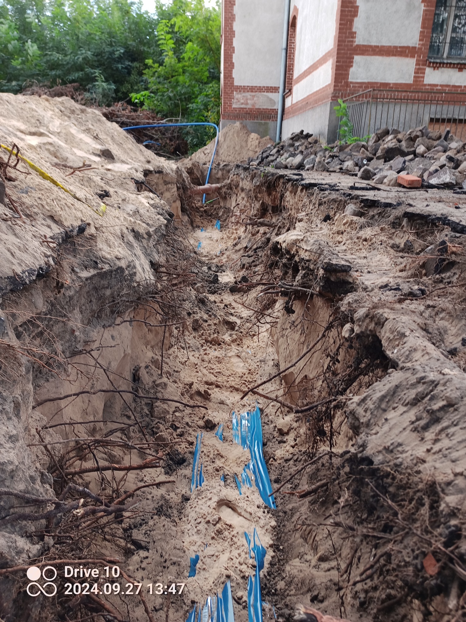 Wykop w ziemi z widocznymi niebieskimi rurami i korzeniami drzew, zabezpieczony folią, obok budynek z ceglaną elewacją.