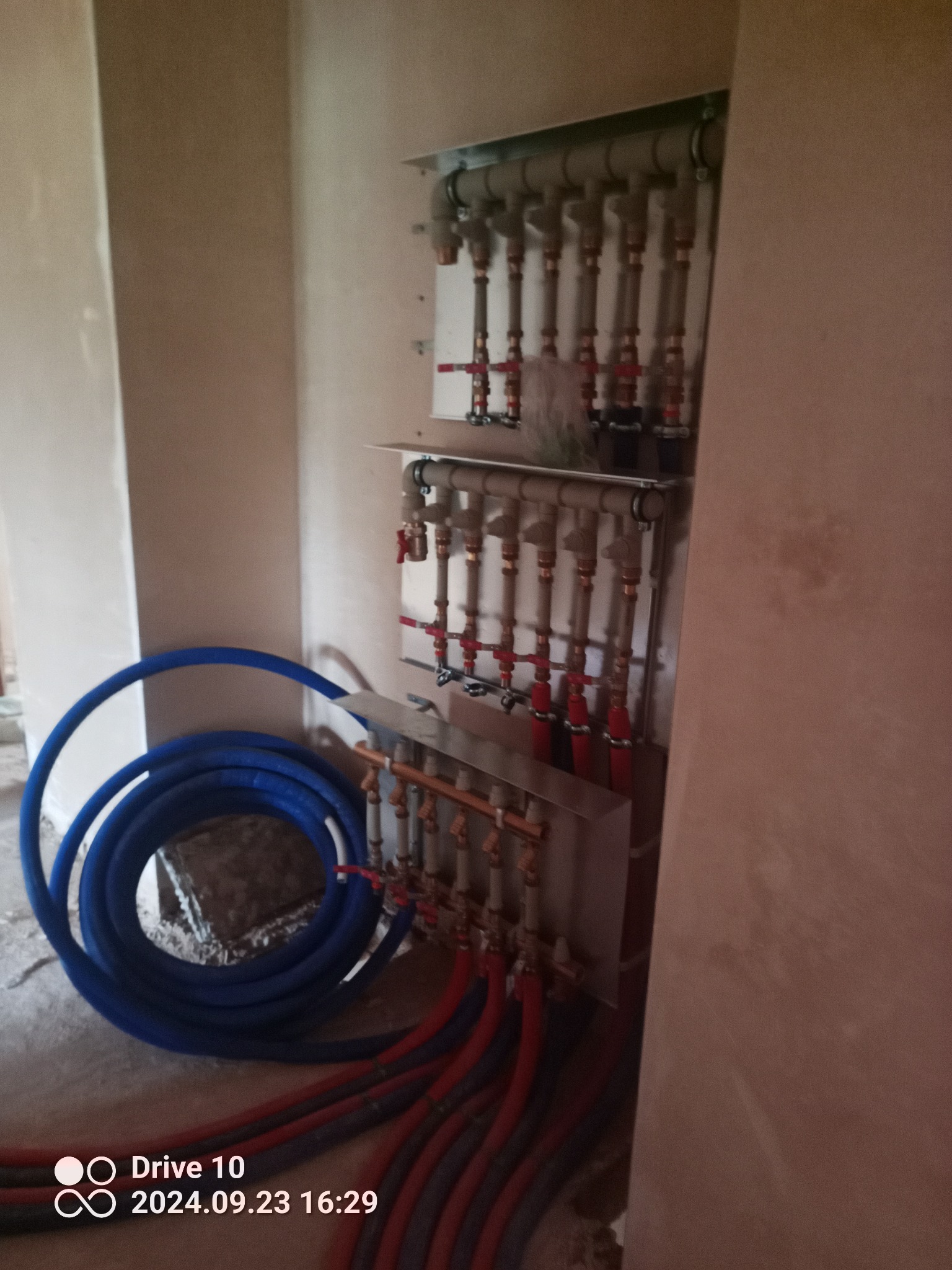 Rozdzielacz ogrzewania podłogowego zamontowany na ścianie w surowym wnętrzu, z widocznymi rurami w kolorze niebieskim i czerwonym, zwiniętymi na podłodze.