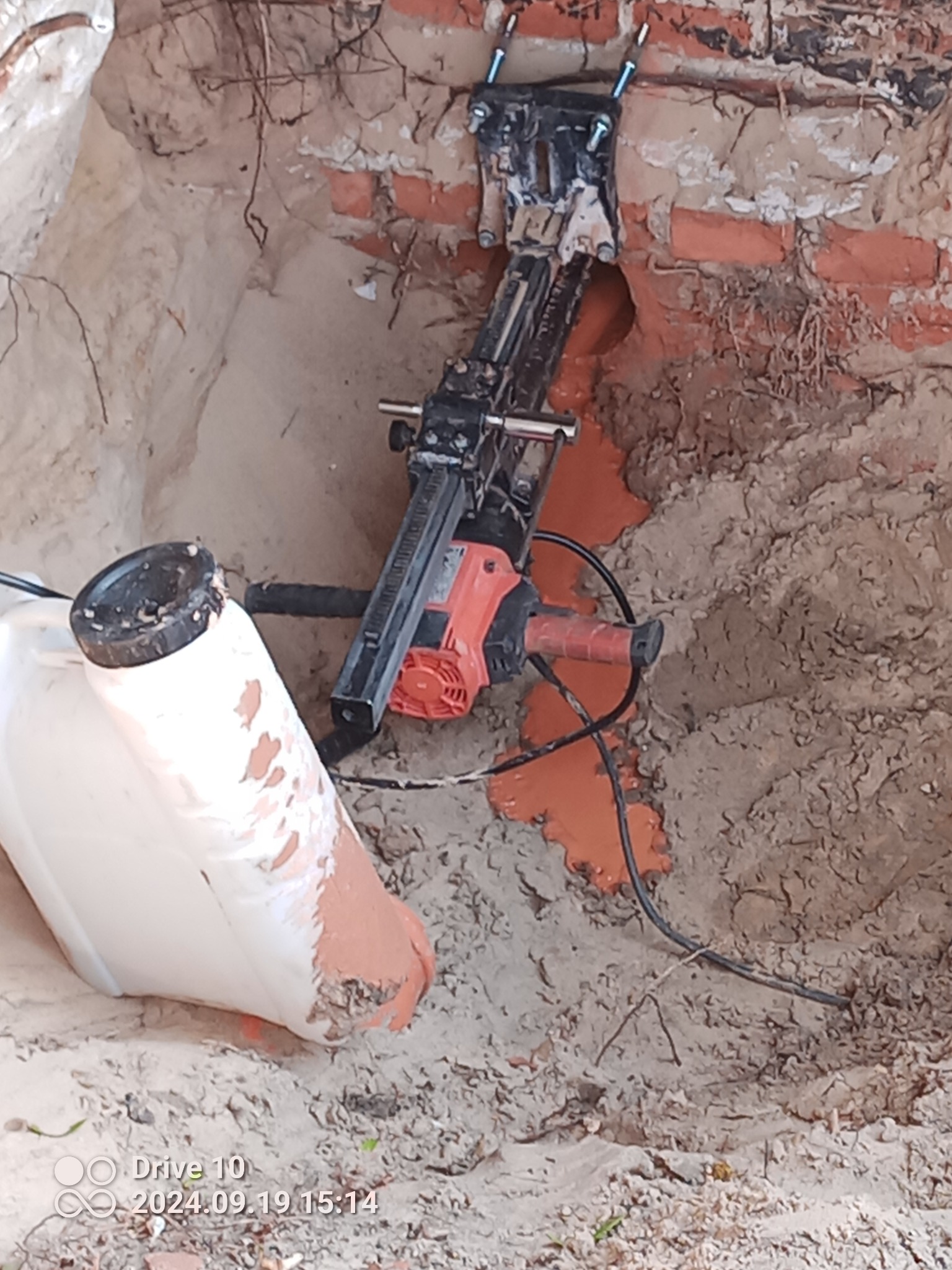 Wiercenie otworu w ceglanej ścianie w wykopie za pomocą wiertnicy diamentowej z podajnikiem wody; widoczny biały kanister z wodą i przewody.