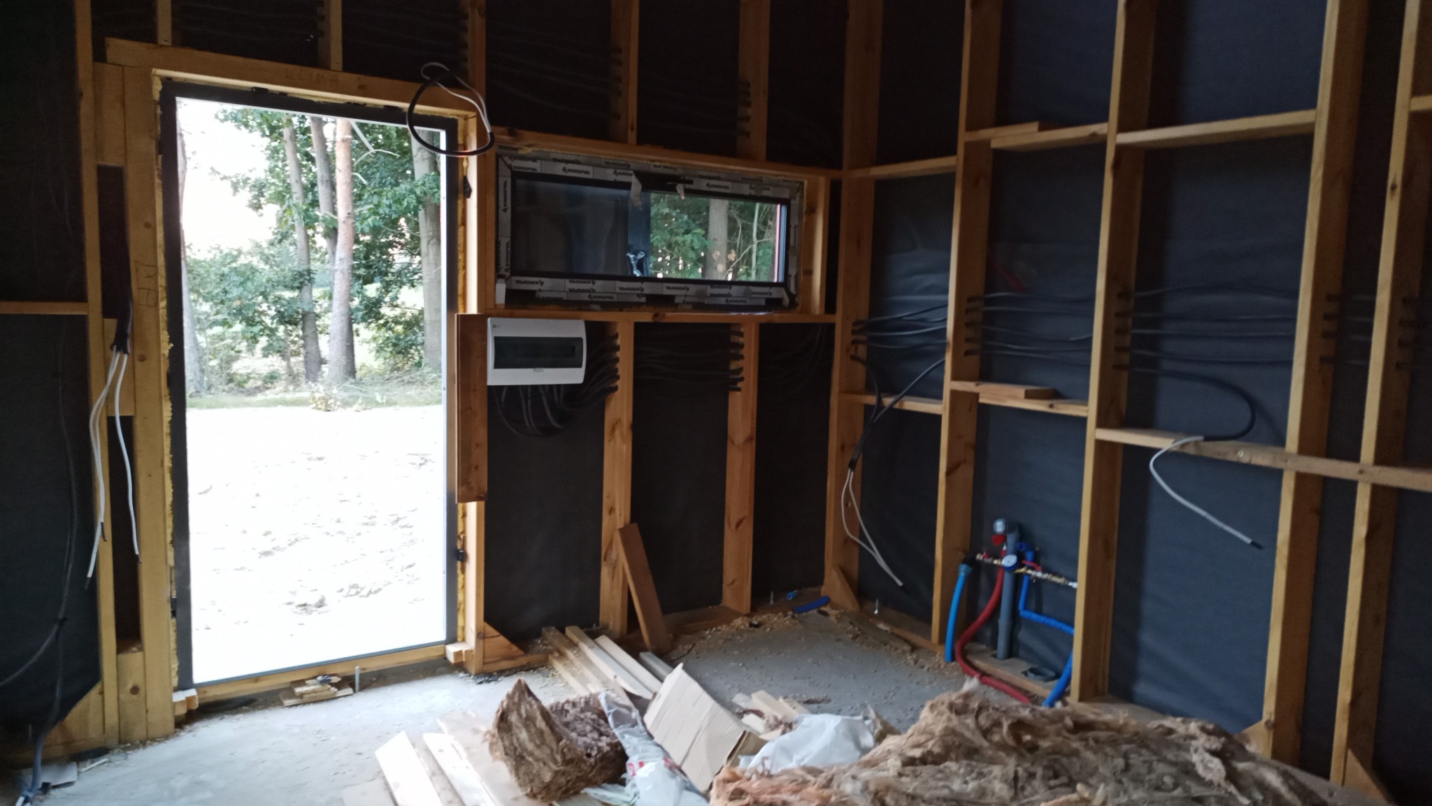 Wnętrze w trakcie budowy z widoczną instalacją elektryczną (rozdzielnia, kable) i hydrauliczną (rury z zaworami) na tle drewnianej konstrukcji szkieletowej i izolacji ścian.
