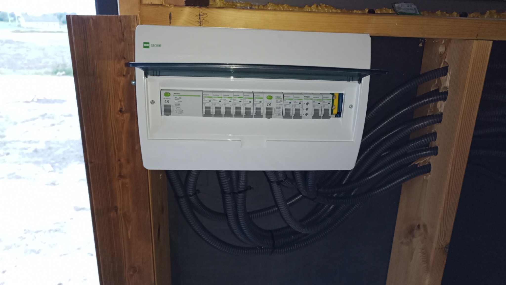 Rozdzielnica elektryczna z otwartą klapką, zamontowana na drewnianej konstrukcji ściany, z widocznymi przewodami w peszlach.