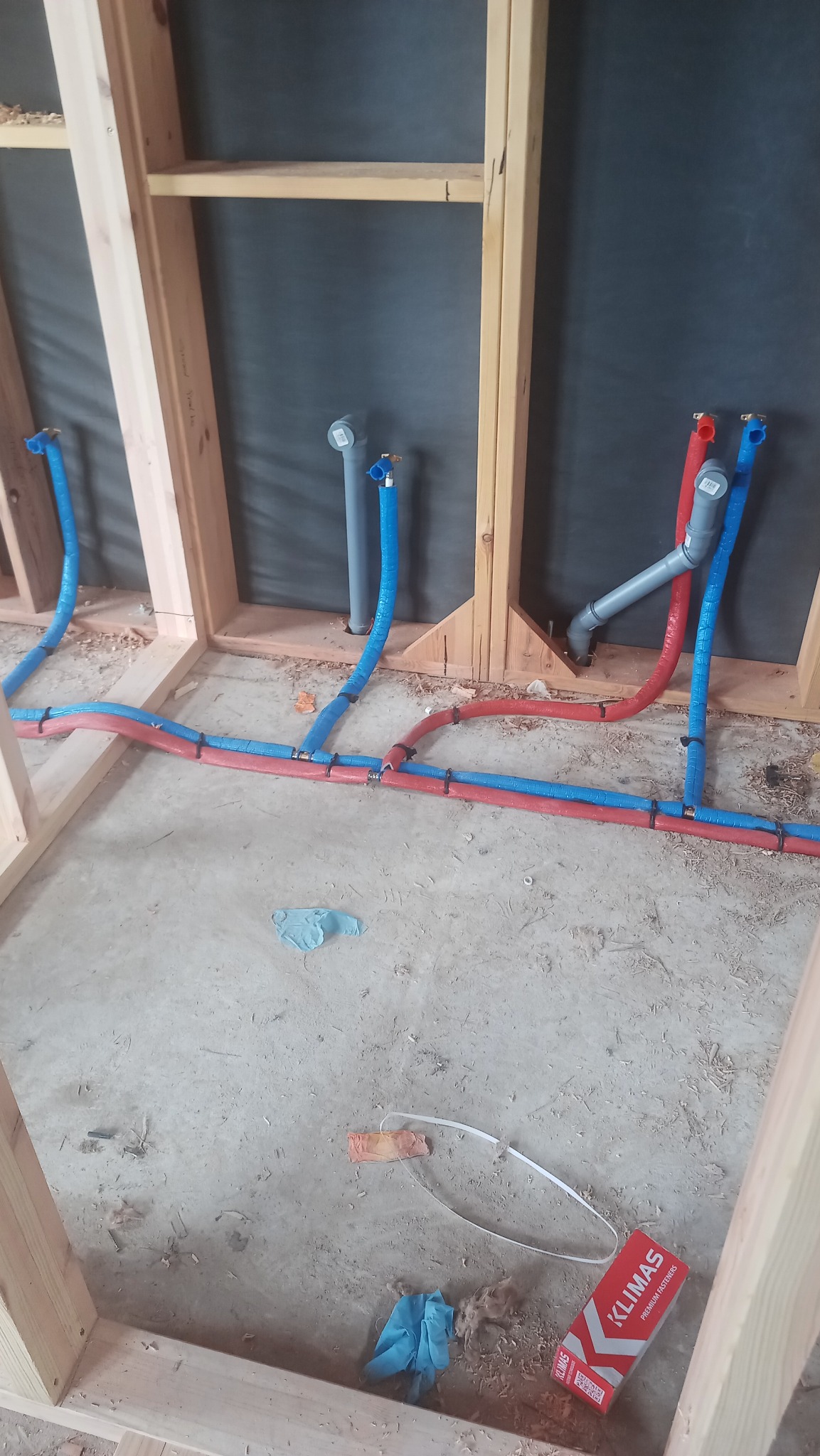 Wnętrze w trakcie budowy z widocznymi, częściowo zamontowanymi rurami instalacji wodnej i kanalizacyjnej w kolorach czerwonym, niebieskim i szarym, przymocowanymi do drewnianej konstrukcji...