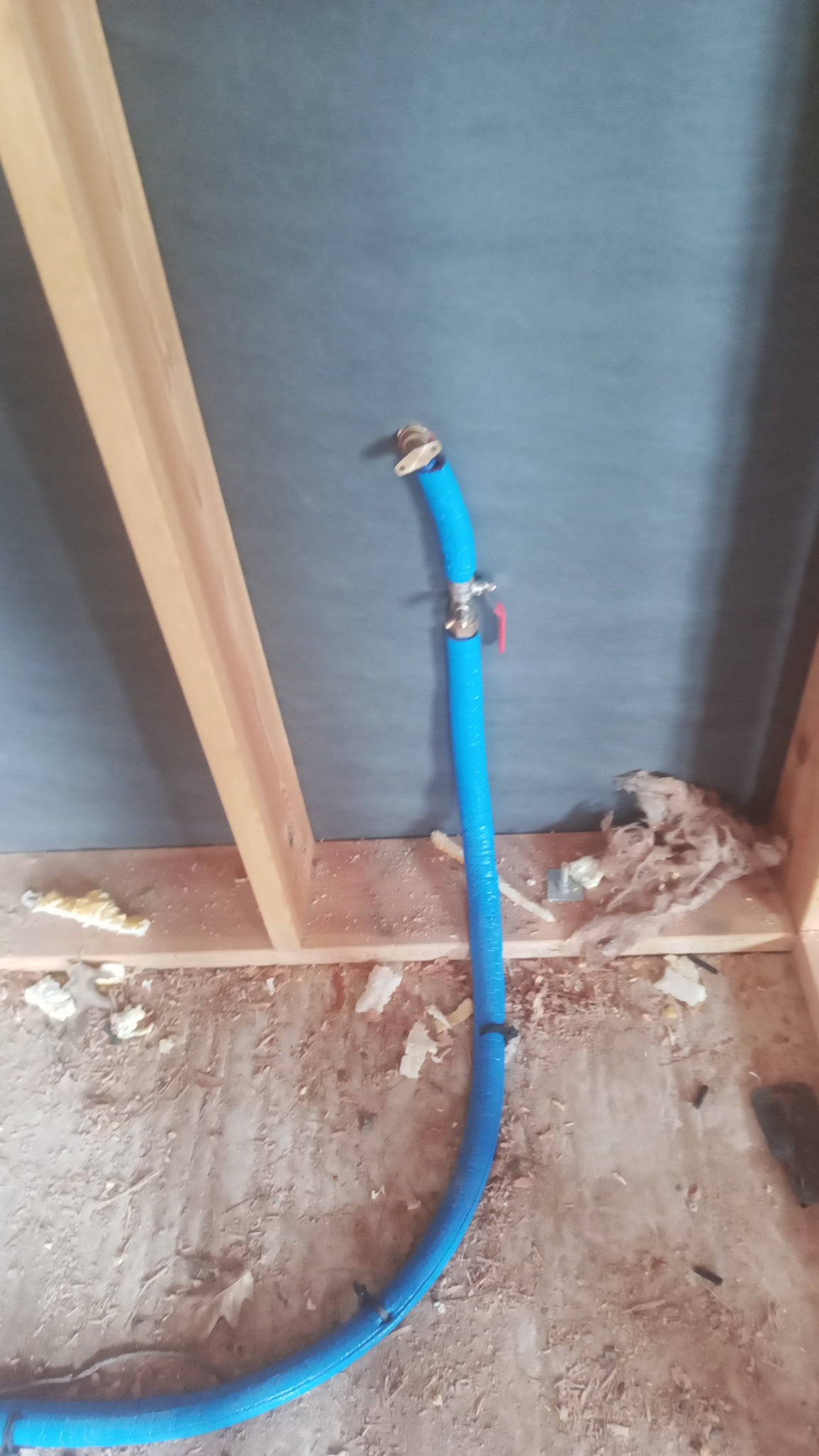Niebieska rura PEX z zaworem kulowym i złączką, prowadzona wzdłuż drewnianej konstrukcji ściany z szarą membraną, widoczne pozostałości izolacji i zabrudzenia na podłodze.