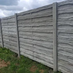 Szary, betonowy płot o fakturze drewna, z widocznymi słupkami i deskami, na tle zieleni i pochmurnego nieba. Solidne ogrodzenie posesji.