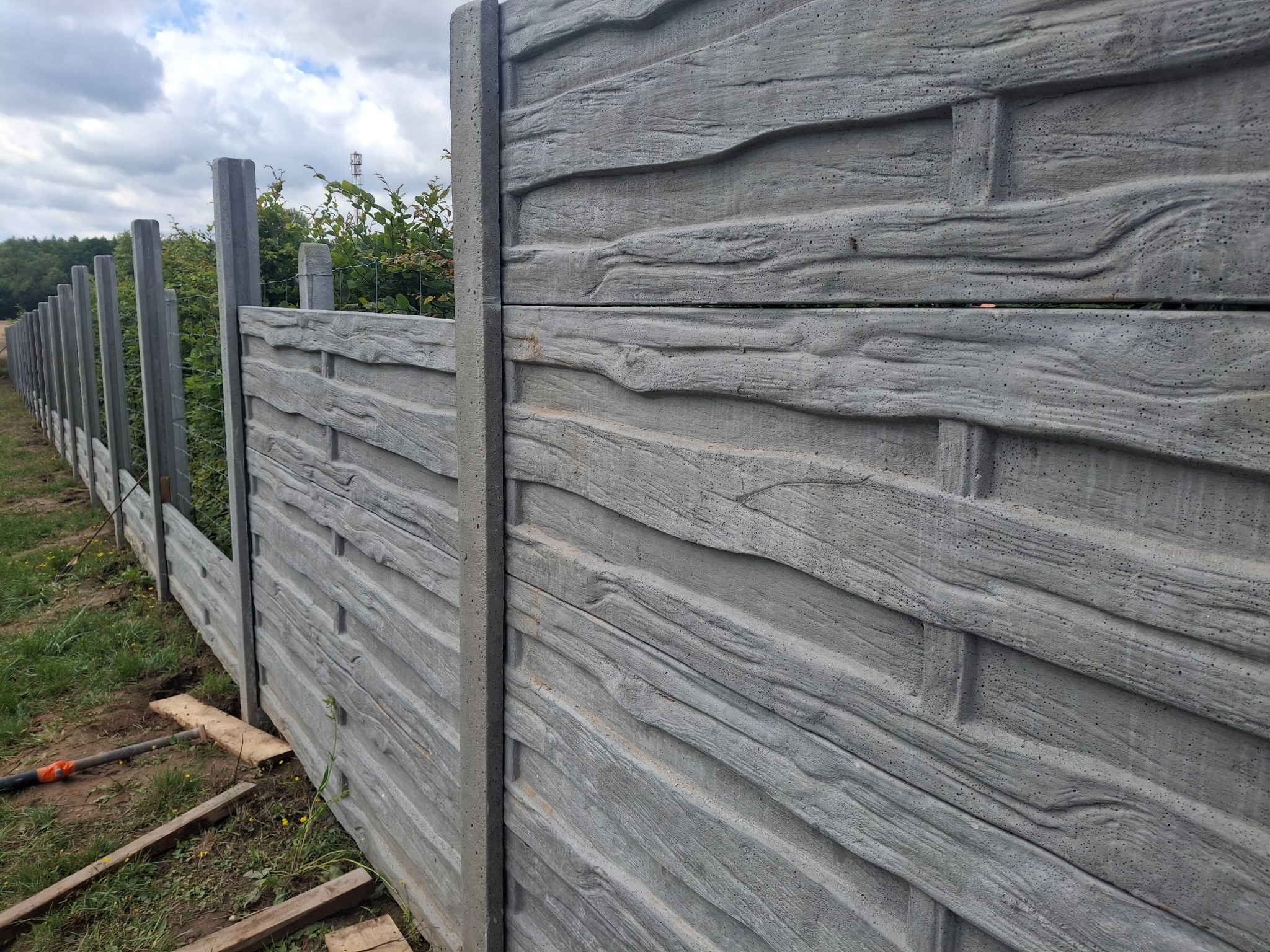 Długie, szare ogrodzenie betonowe z fakturą drewna, z widocznymi słupkami i ziemią u podstawy, rozciągające się wzdłuż zieleni i drzew.