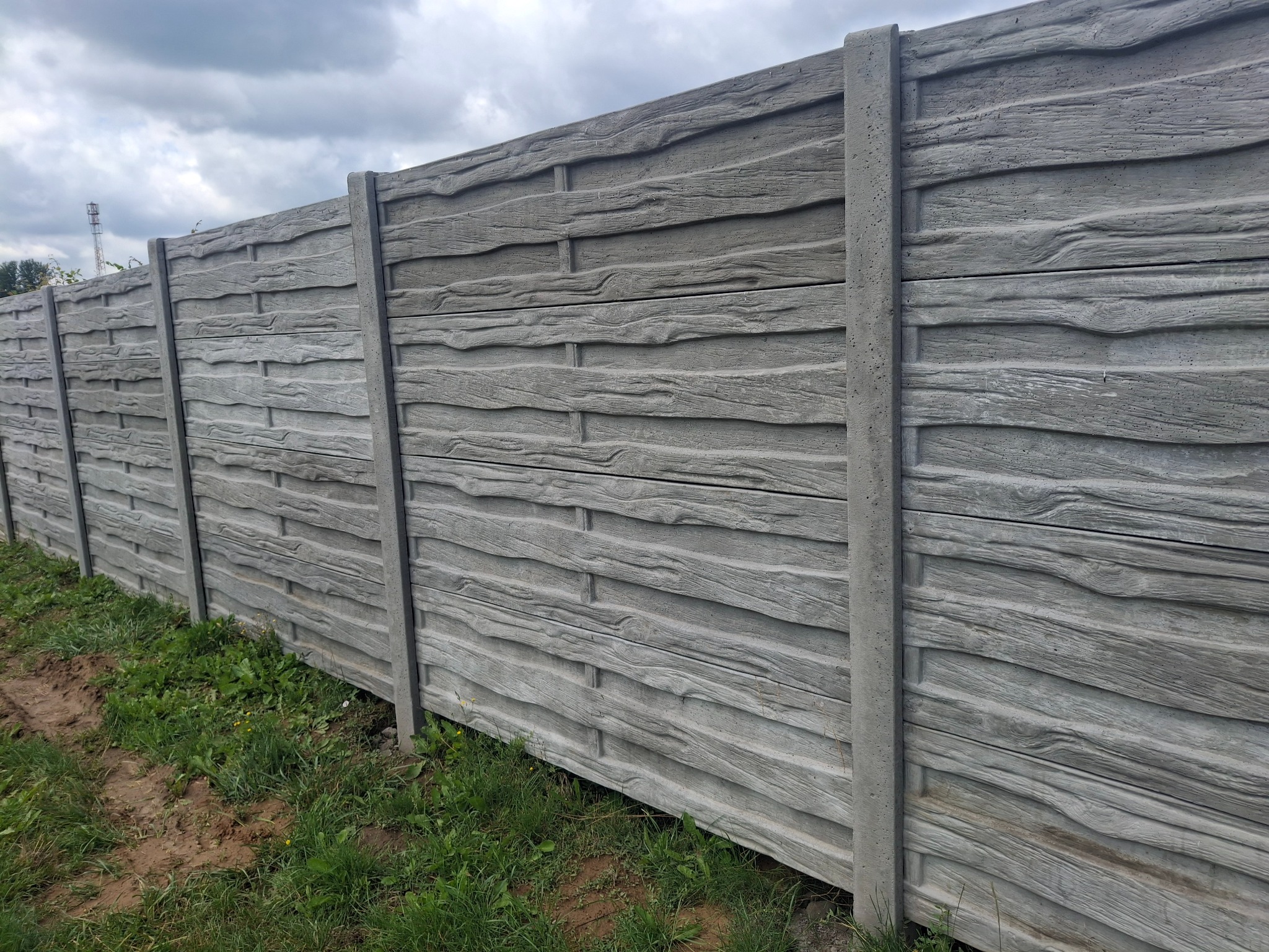 Szary, betonowy płot o fakturze drewna, z widocznymi słupkami i deskami, na tle zieleni i pochmurnego nieba. Solidne ogrodzenie posesji.