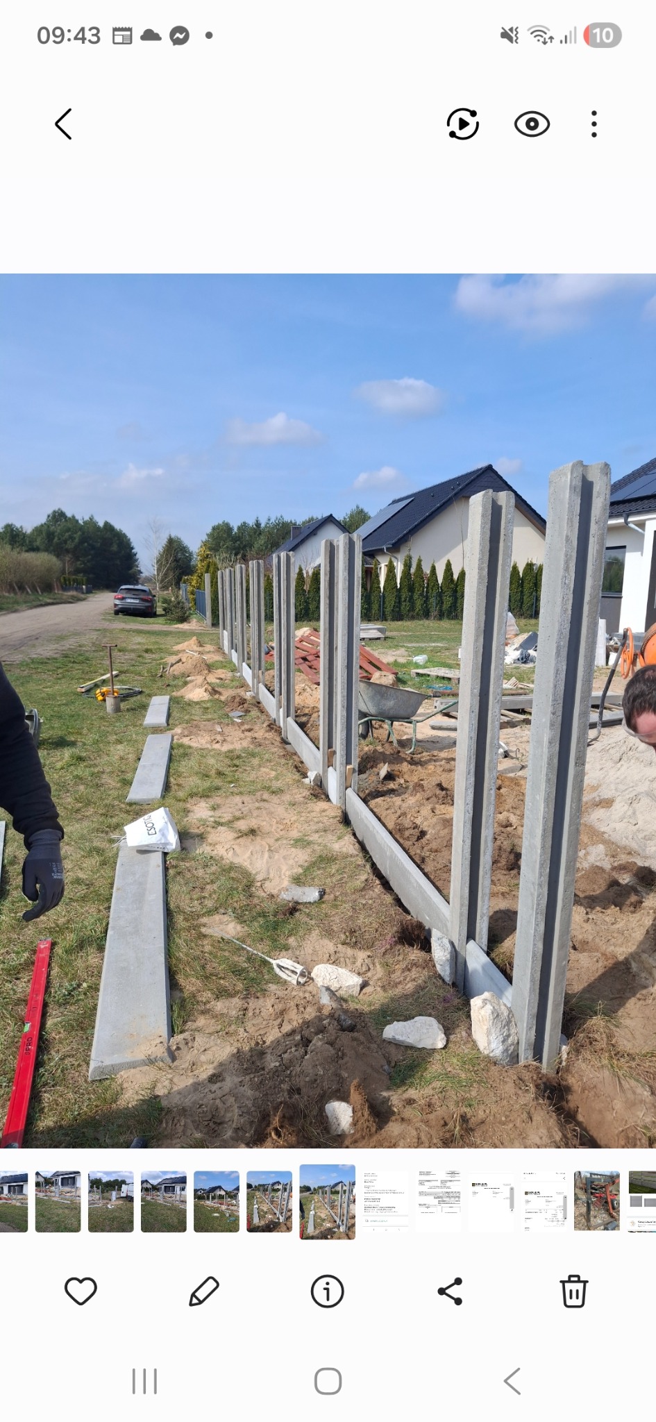 Montaż betonowego ogrodzenia panelowego: ustawione słupki i podmurówka na działce z widokiem na domy. Widoczne narzędzia i materiały budowlane.