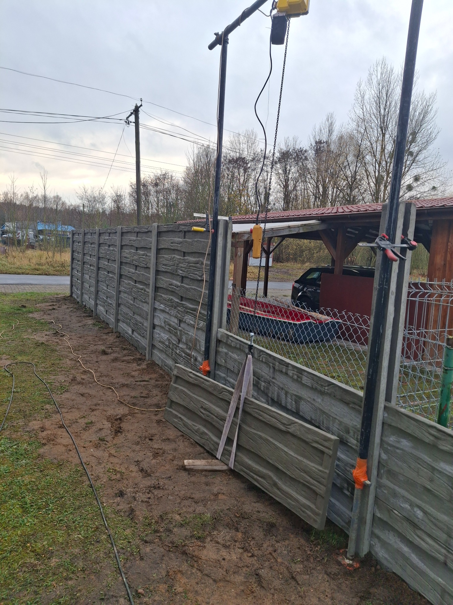 Montaż ogrodzenia betonowego z użyciem dźwigu. Widoczne panele o fakturze drewna i elementy montażowe. Tło stanowią drzewa i zabudowania.