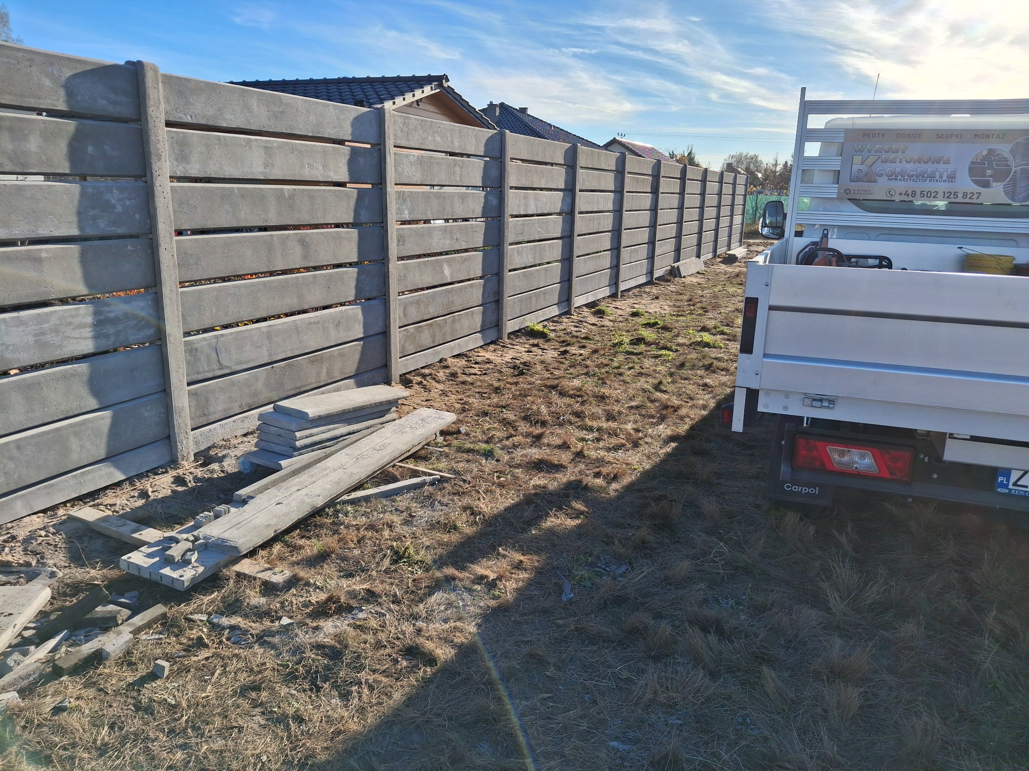 Długi, szary, betonowy płot panelowy z poziomymi szczelinami, częściowo zamontowany, z samochodem dostawczym w tle i stertą płyt na ziemi.