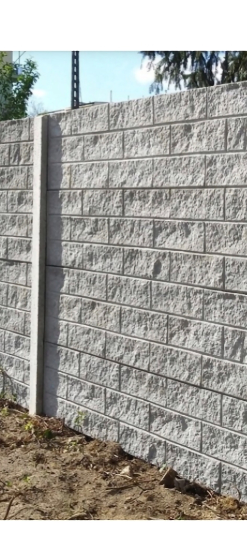 Szary, teksturowany mur z betonowych bloczków, tworzący ogrodzenie. Widoczny słupek ogrodzeniowy i fragment ziemi u podstawy. Tło rozmyte, z elementami zieleni.