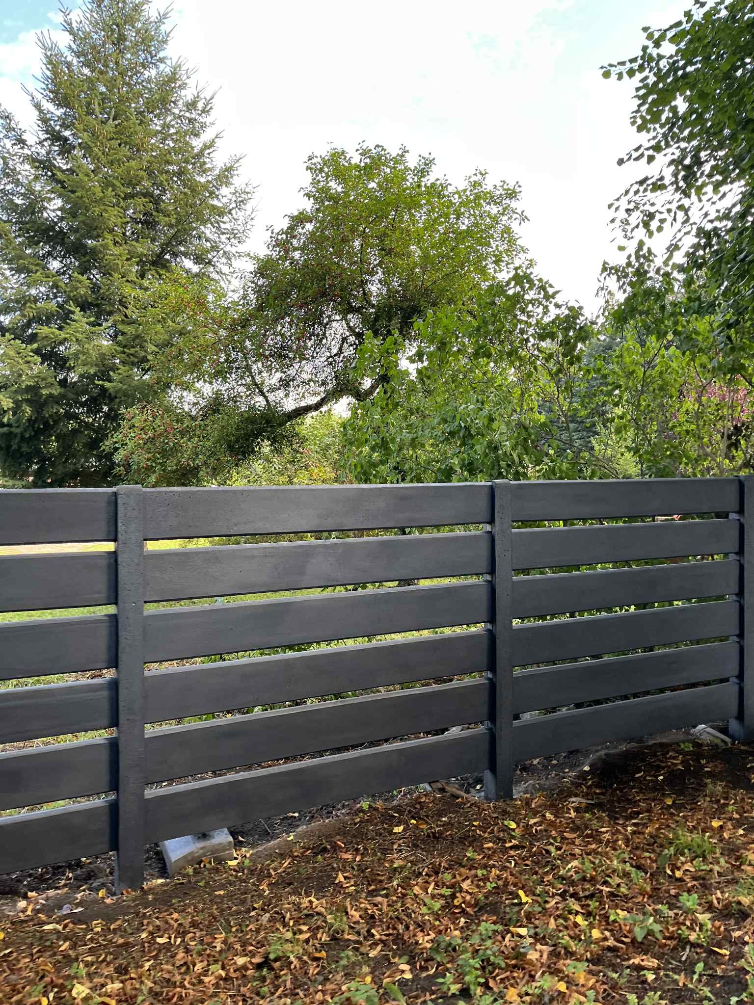 Nowoczesne, szare ogrodzenie panelowe z betonu, z poziomymi szczelinami, na tle zieleni drzew. Widoczne betonowe podstawy słupków.