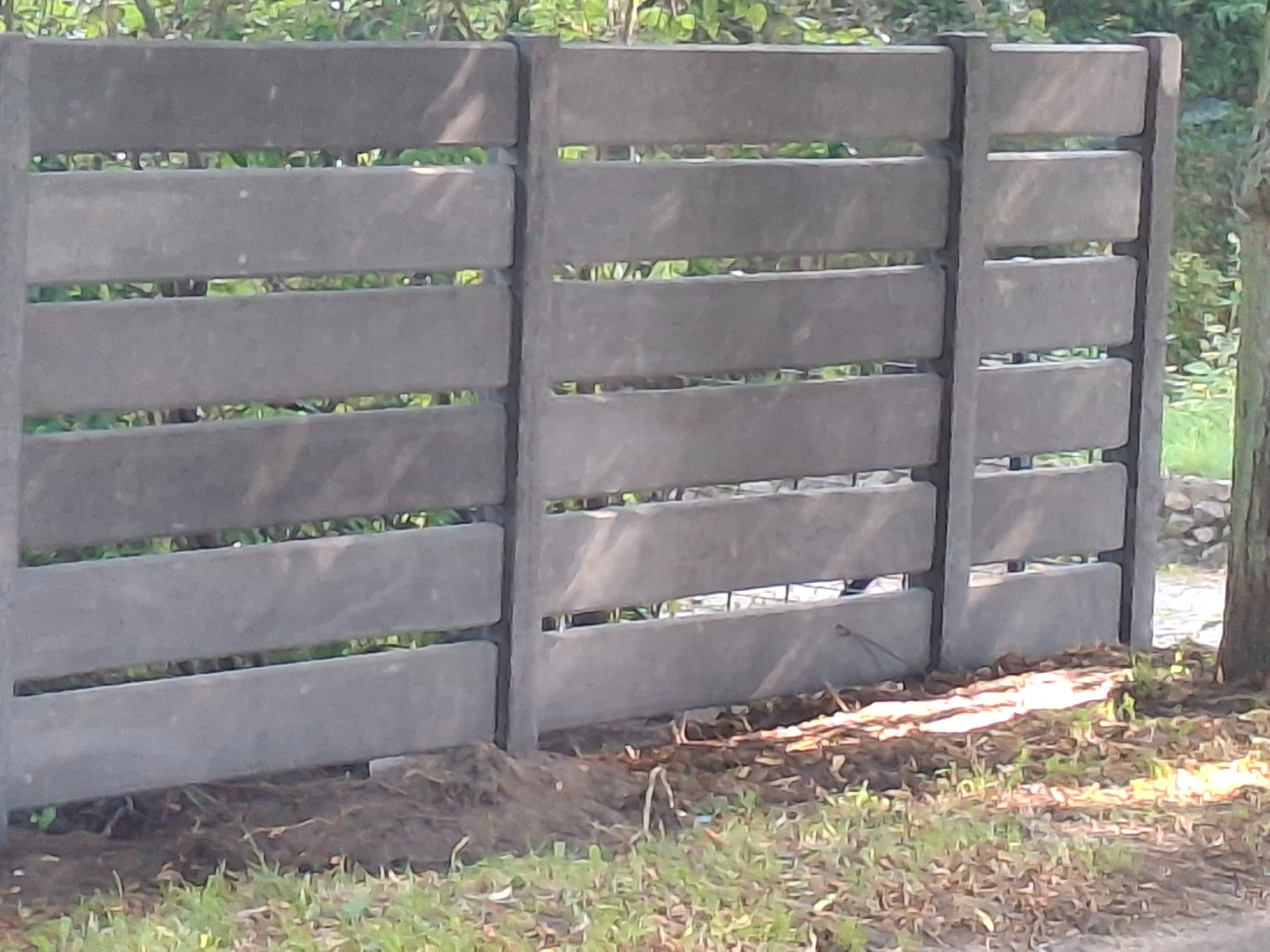 Nowoczesne, szare ogrodzenie betonowe z poziomymi panelami, widoczne na tle zieleni. Minimalistyczny design, regularne odstępy między elementami.