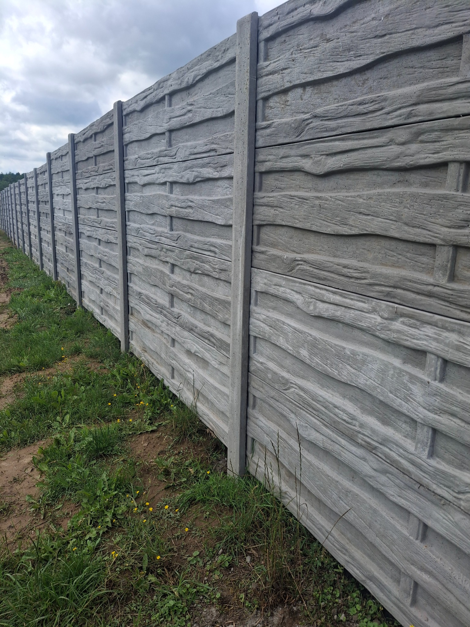 Długi, szary, betonowy płot z fakturą drewna, oddzielający teren zielony. Widok z boku, z perspektywą zbieżną w dal. Pochmurne niebo w tle.