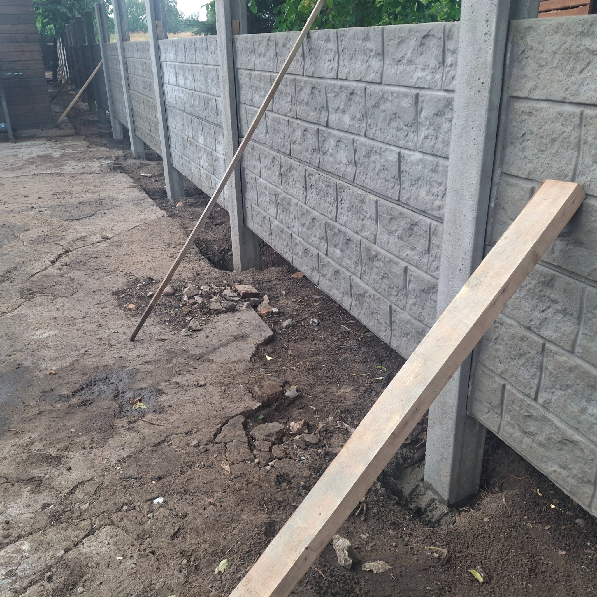 Fragment montażu ogrodzenia betonowego z teksturą cegły. Widoczne słupki i panele w trakcie instalacji, z ziemią i drewnianymi elementami pomocniczymi.