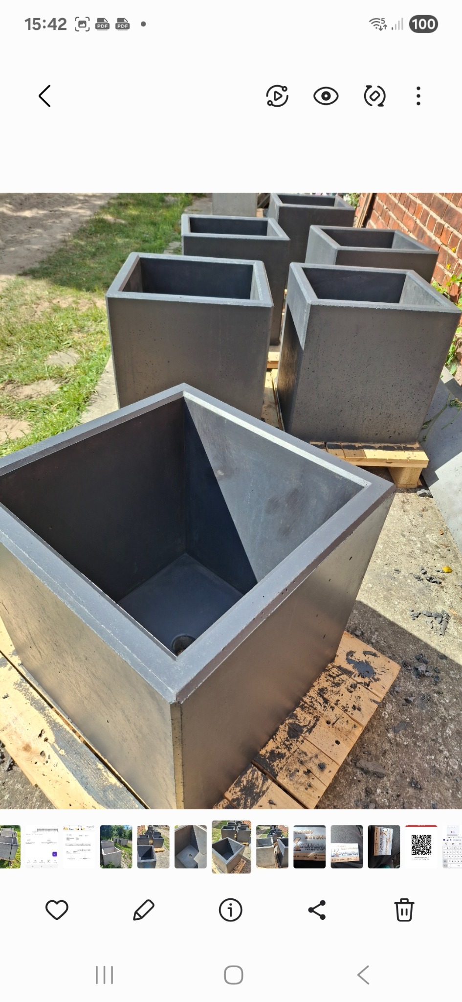 Szare, betonowe donice w kształcie sześcianów, ustawione na palecie. Widoczne detale faktury betonu i otwór drenażowy w jednej z donic. Tło z ceglanym murem.