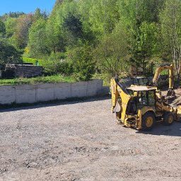 CT SZKOLENIA SPÓŁKA Z OGRANICZONĄ ODPOWIEDZIALNOŚCIĄ - Żółta koparko-ładowarka Caterpillar na placu budowy, z widocznym stosem ziemi i betonowym ogrodzeniem w tle, w słoneczny dzień.