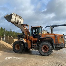 CT SZKOLENIA SPÓŁKA Z OGRANICZONĄ ODPOWIEDZIALNOŚCIĄ - Pomarańczowa ładowarka jednonaczyniowa Doosan DL400 z podniesioną łyżką wypełnioną piaskiem, widoczny operator w kabinie, w tle betonowe bloki i drzewa, na ziemi ślady opon.