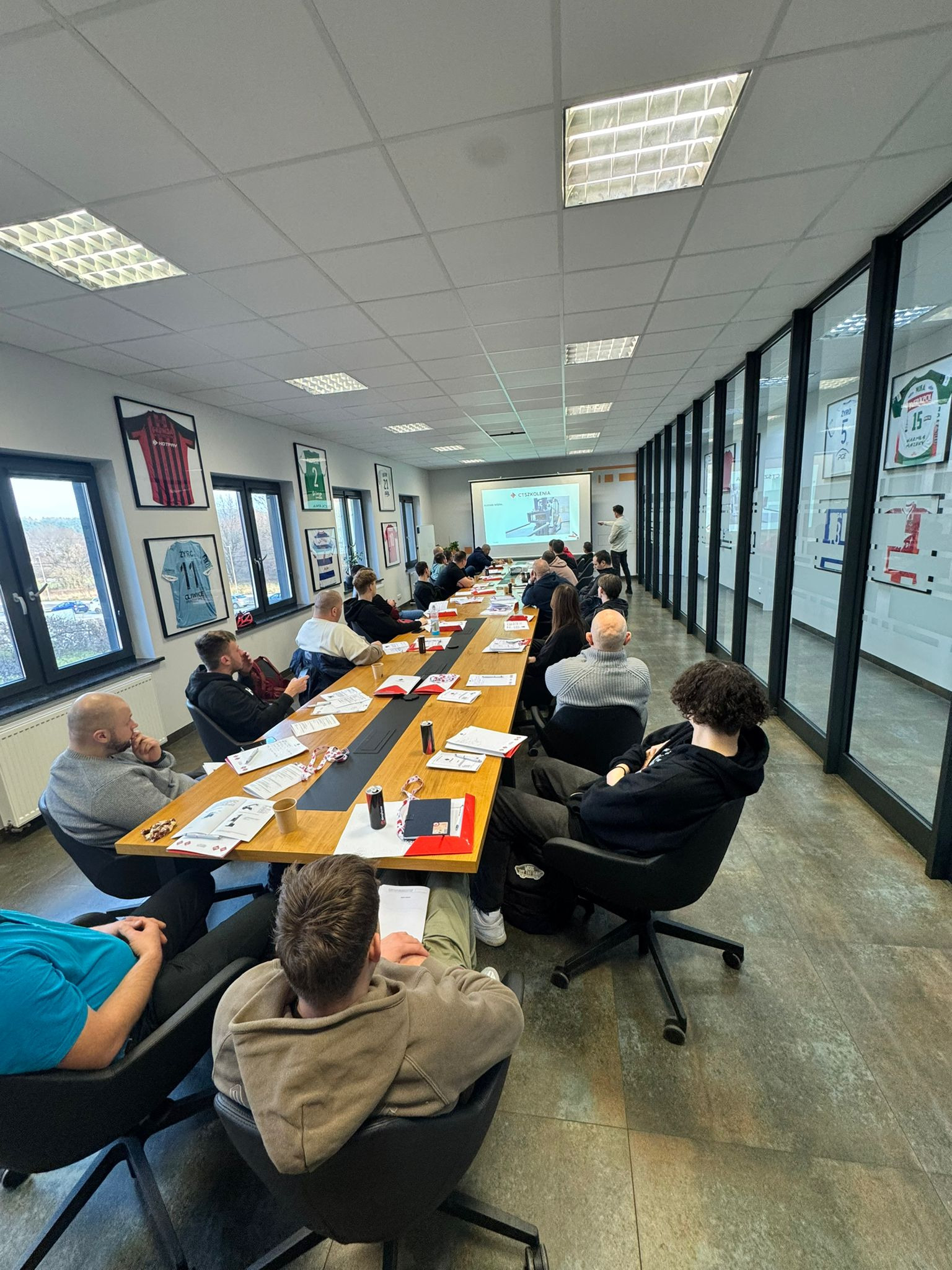 Sala szkoleniowa z uczestnikami siedzącymi przy długim stole, prowadzący prezentuje slajd z wózkiem widłowym, na ścianach wiszą oprawione koszulki sportowe.
