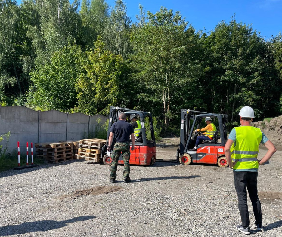 Instruktorzy i kursanci podczas zajęć praktycznych z obsługi wózków widłowych na placu manewrowym z paletami, betonowym ogrodzeniem i drzewami w tle.