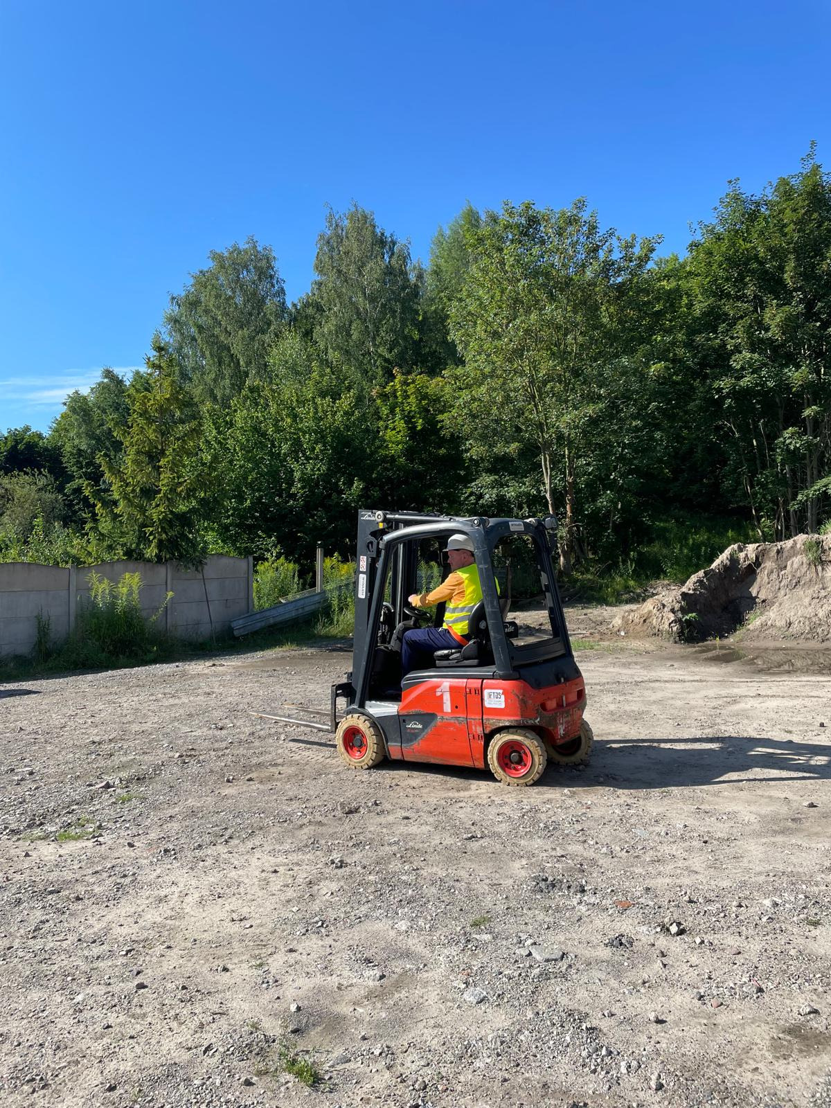 Operator w kasku i kamizelce odblaskowej prowadzi pomarańczowo-czarny wózek widłowy Linde po żwirowym terenie, z widocznym lasem w tle pod błękitnym niebem.