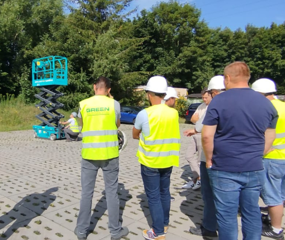 Grupa osób w kaskach i kamizelkach odblaskowych obserwuje demonstrację podnośnika nożycowego na placu manewrowym podczas kursu zawodowego.