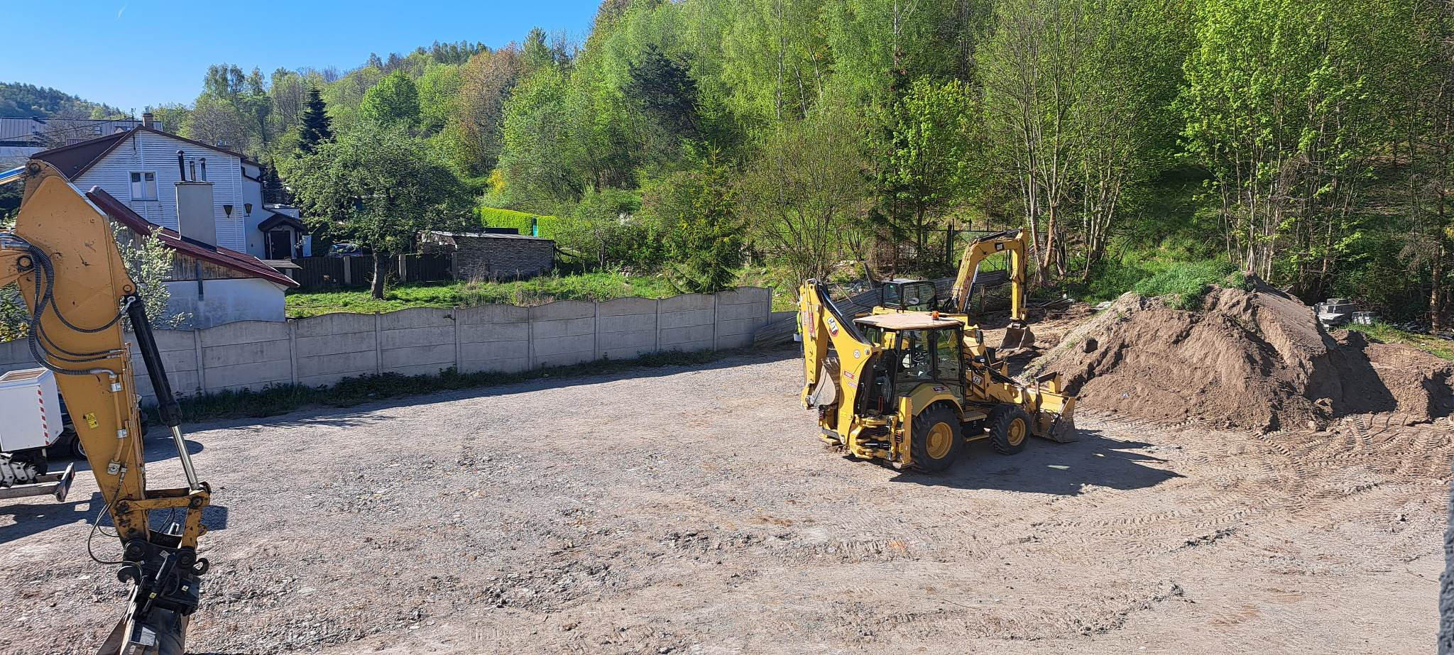 Żółta koparko-ładowarka Caterpillar na placu budowy, z widocznym stosem ziemi i betonowym ogrodzeniem w tle, w słoneczny dzień.