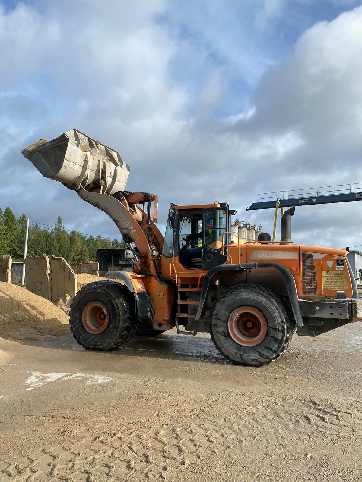 Pomarańczowa ładowarka jednonaczyniowa Doosan DL400 z podniesioną łyżką wypełnioną piaskiem, widoczny operator w kabinie, w tle betonowe bloki i drzewa, na ziemi ślady opon.