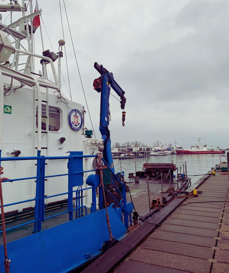 Widok na pokład statku Urzędu Morskiego w Gdyni z dźwigiem, cumującego przy drewnianym nabrzeżu, w tle inne jednostki pływające i zabudowania portowe.