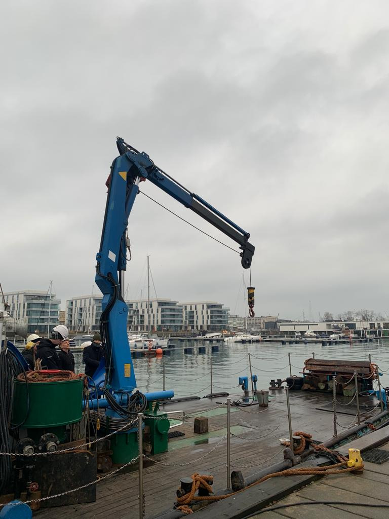Niebieski dźwig na platformie z widokiem na port i budynki mieszkalne w tle, grupa osób w kaskach obserwuje pracę dźwigu.