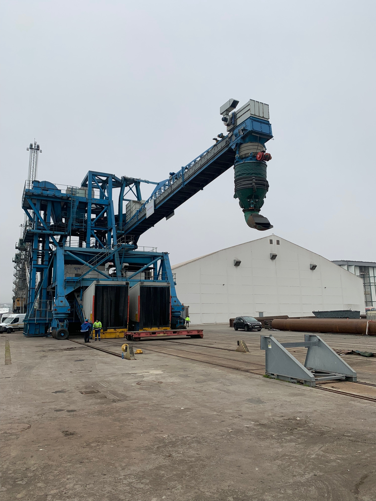 Niebieski suwnicowy żuraw portowy z podwieszonym zasobnikiem ładunkowym, pracownicy w odblaskowych kamizelkach nadzorują załadunek do kontenerów, tło stanowi magazyn i pochmurne niebo.