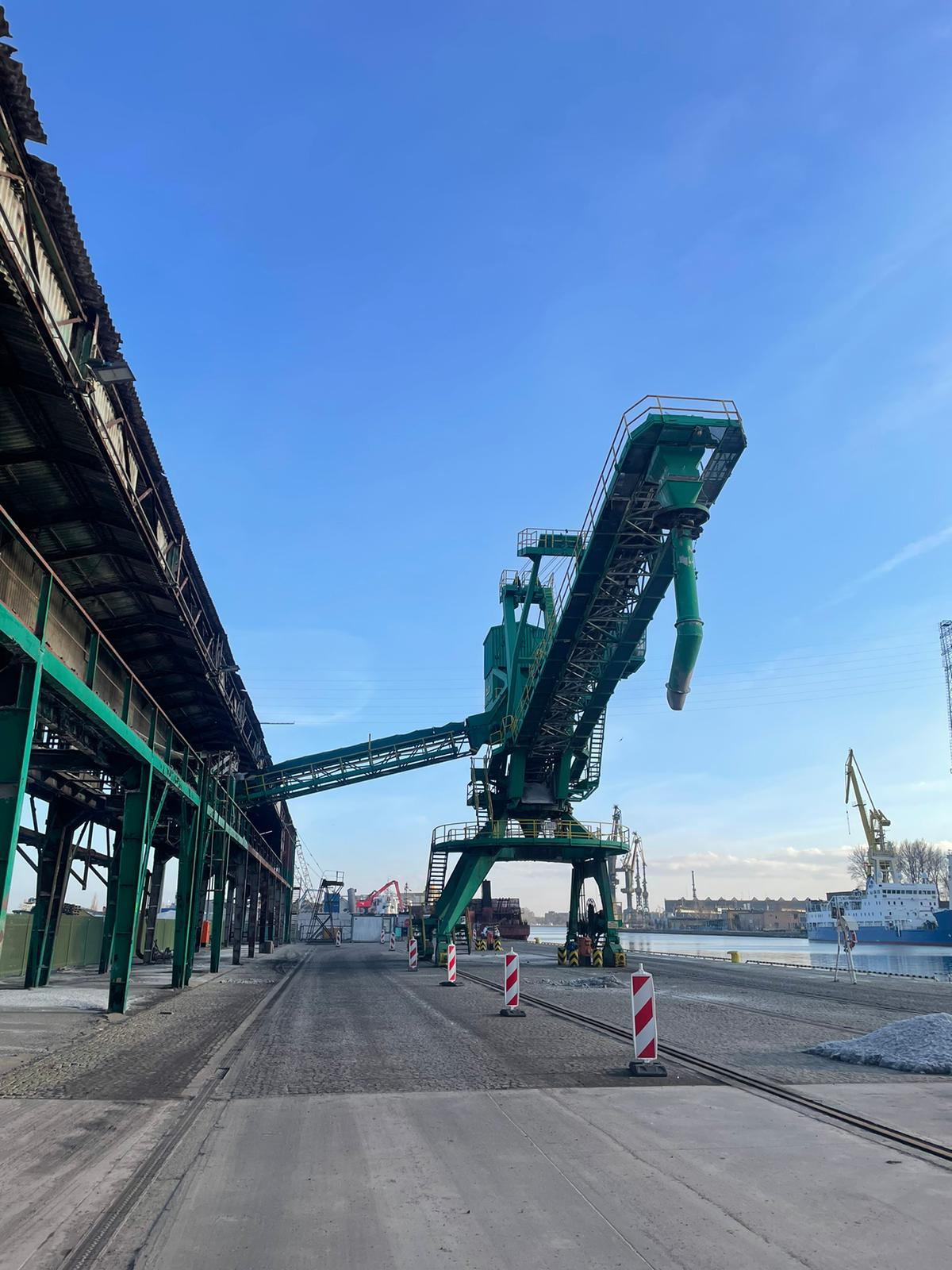 Zielony dźwig portowy z podajnikiem taśmowym na tle błękitnego nieba i nabrzeża z innymi dźwigami i statkami.