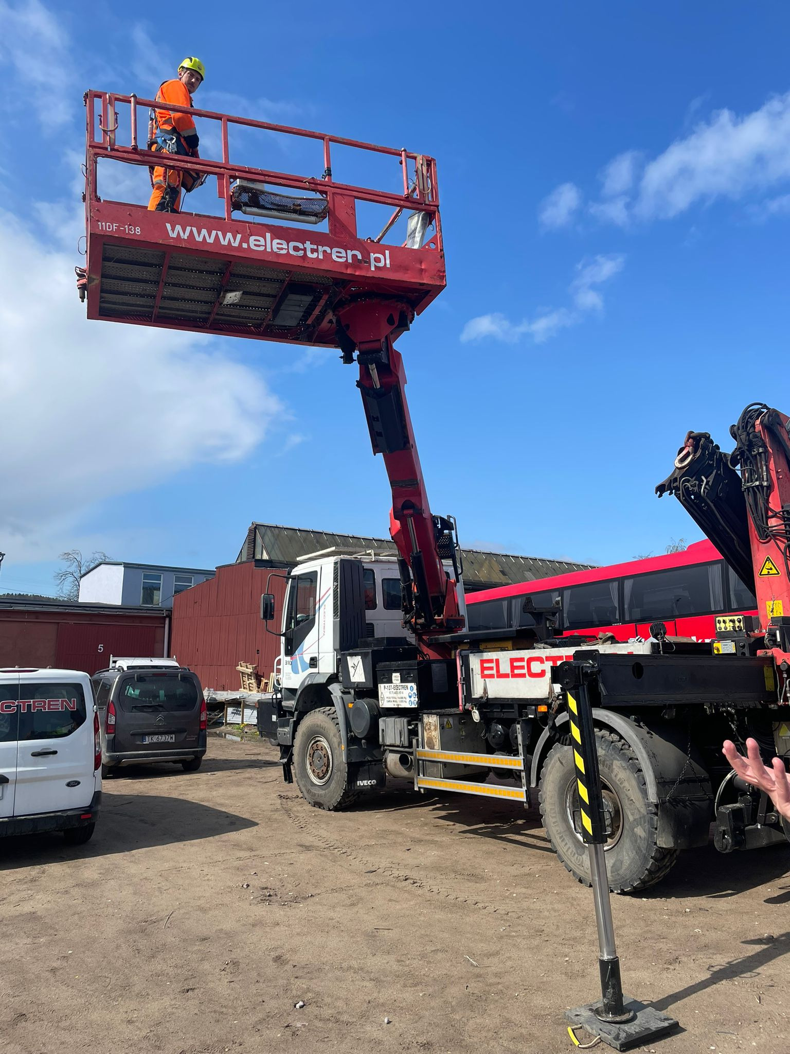 Pracownik w pomarańczowym kombinezonie i żółtym kasku na podnośniku koszowym firmy Electren, na tle błękitnego nieba z chmurami.