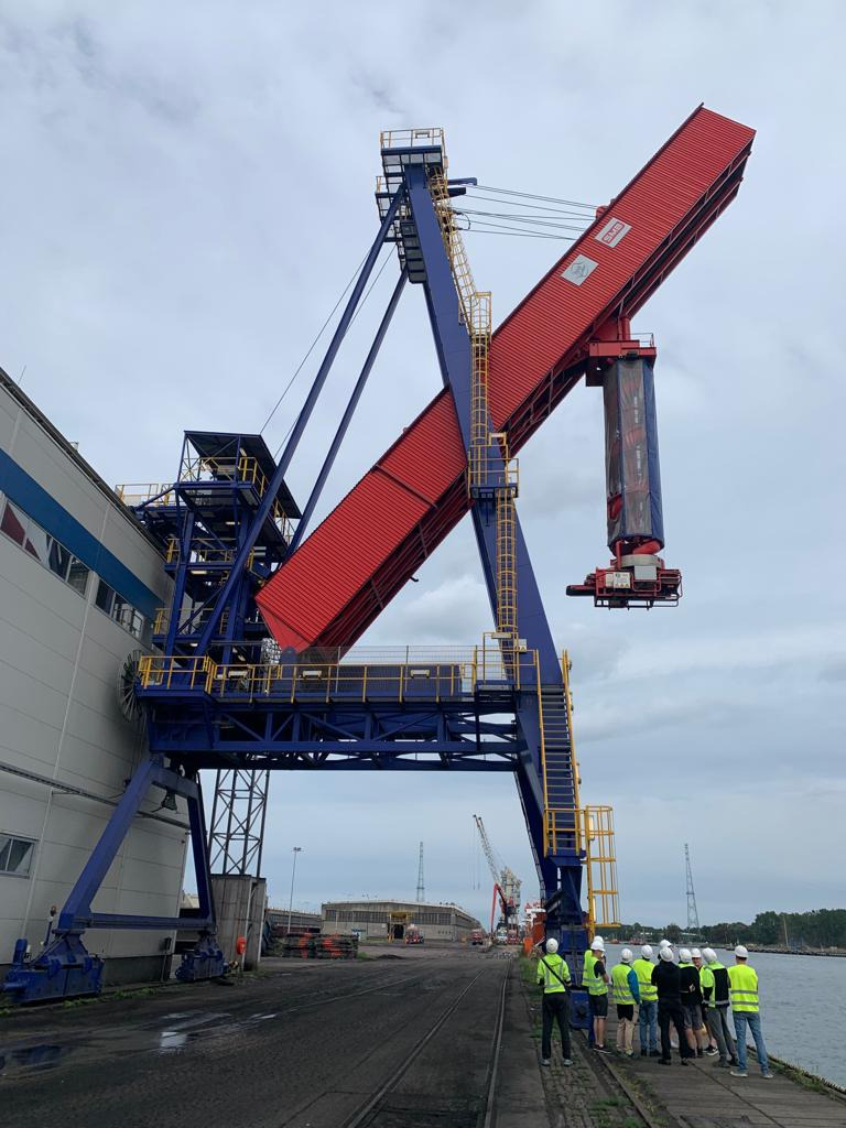 Wysoki suwnicowy dźwig portowy w kolorach niebieskim i czerwonym, z grupą osób w kaskach i kamizelkach odblaskowych u jego podstawy, obserwujących pracę urządzenia.