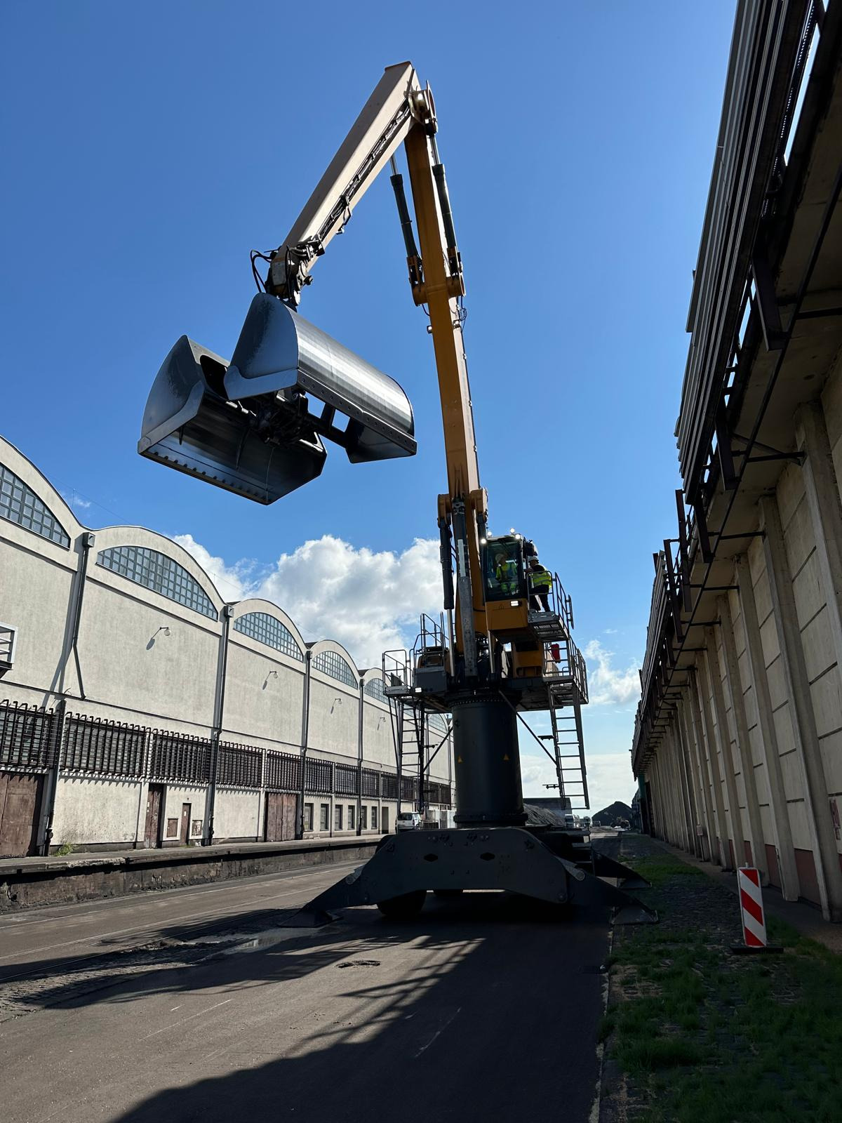 Żuraw portowy z operatorem, widok z dołu na tle błękitnego nieba i budynków portowych.