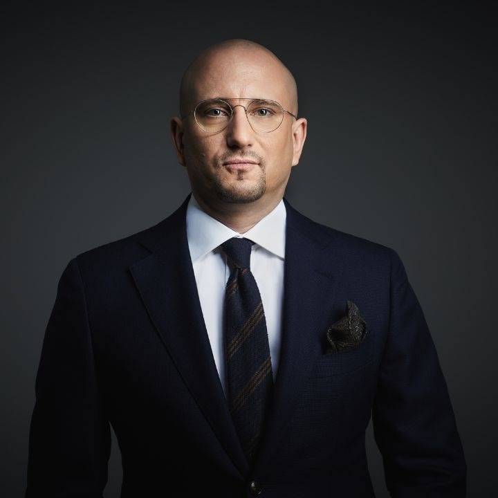 Portret łysiejącego mężczyzny w okularach i granatowym garniturze z poszetką, na szarym tle.