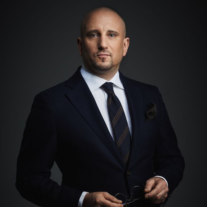 Portret mężczyzny w granatowym garniturze, białej koszuli i brązowo-granatowym krawacie, trzymającego okulary w dłoniach na tle ciemnoszarego tła.