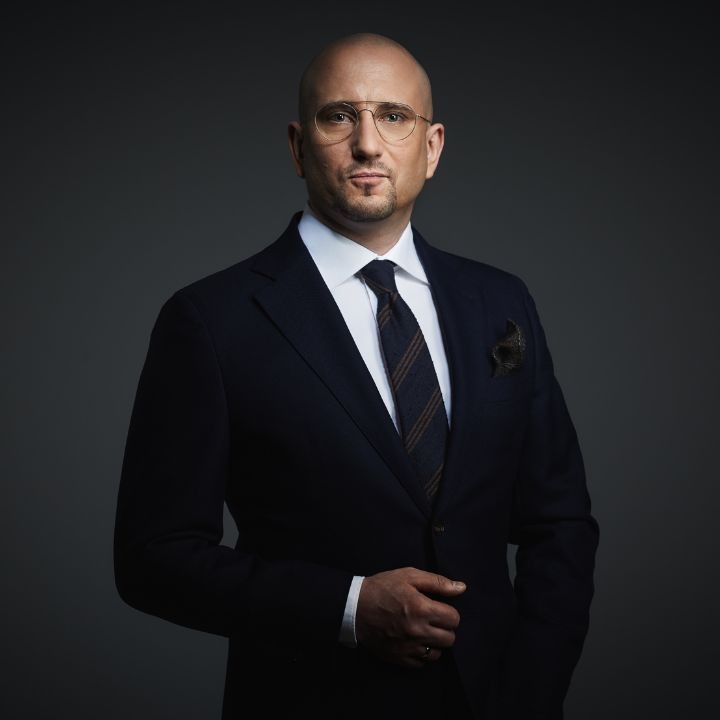 Portret mężczyzny w ciemnym garniturze, białej koszuli i krawacie w paski, z poszetką w kieszonce, na ciemnoszarym tle.