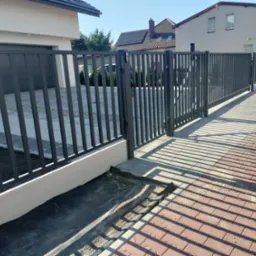 Nowoczesna, grafitowa brama przesuwna z pionowych profili metalowych, z widocznymi panelami fotowoltaicznymi na dachu domu w tle, rzucająca długie cienie na chodnik z kostki.