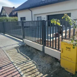 Nowoczesne ogrodzenie metalowe w kolorze grafitowym, z pionowymi prętami, otaczające posesję z domem jednorodzinnym; widoczna skrzynka gazowa w kolorze żółtym i fragment chodnika z kostki.